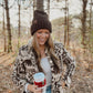 Woman in a patterned jacket and beanie holding a coffee cup in a forest setting