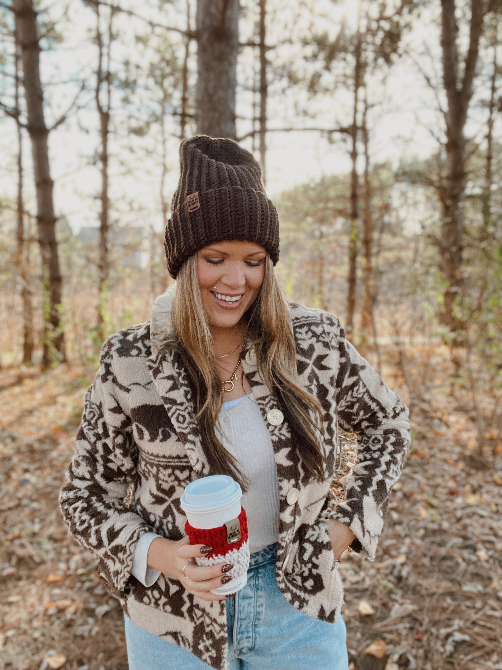 Woman in a patterned jacket and beanie holding a coffee cup in a forest setting